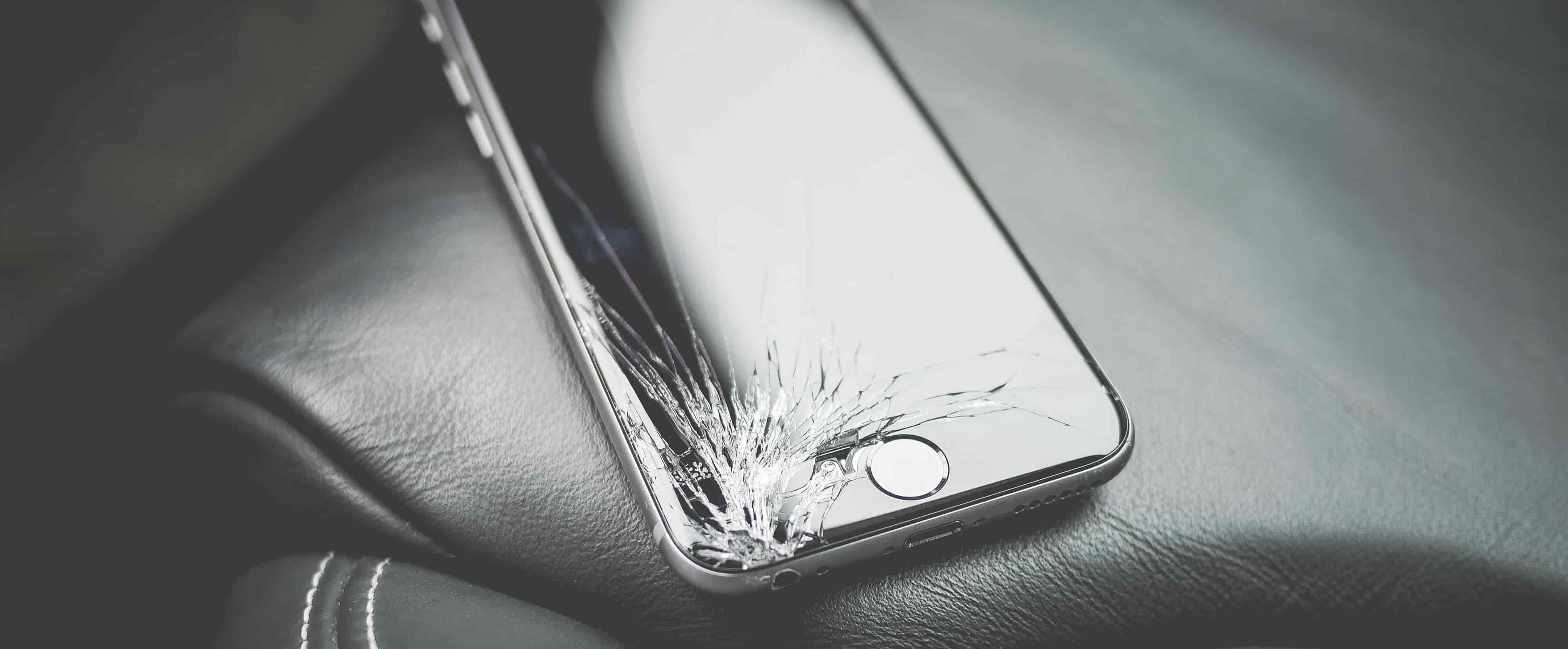 A black iPhone with a cracked screen sitting on a black leather car seat